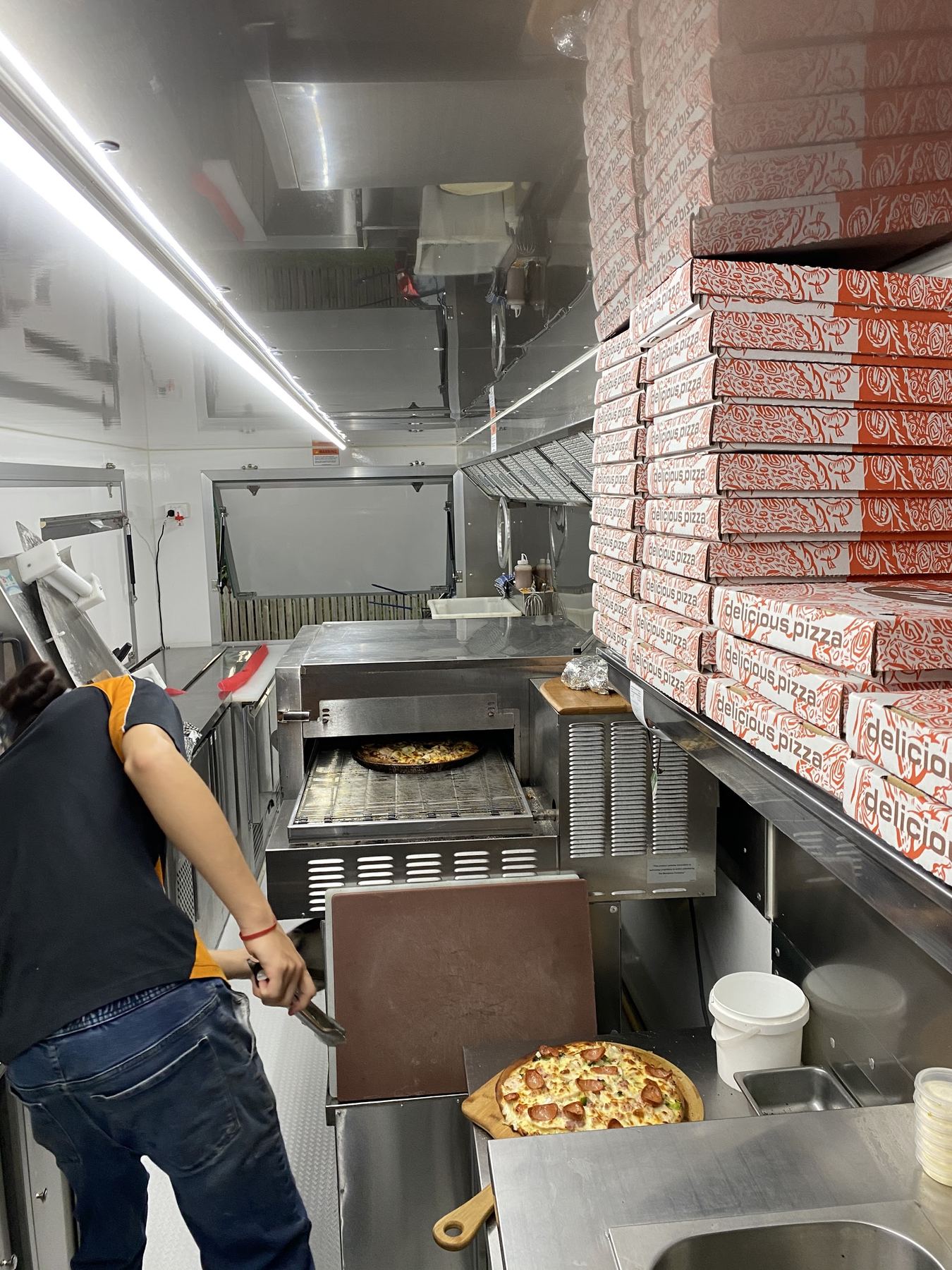 A pizza coming out of the conveyor oven inside the Blaze Pizzas trailer, with boxes stacked ready to go